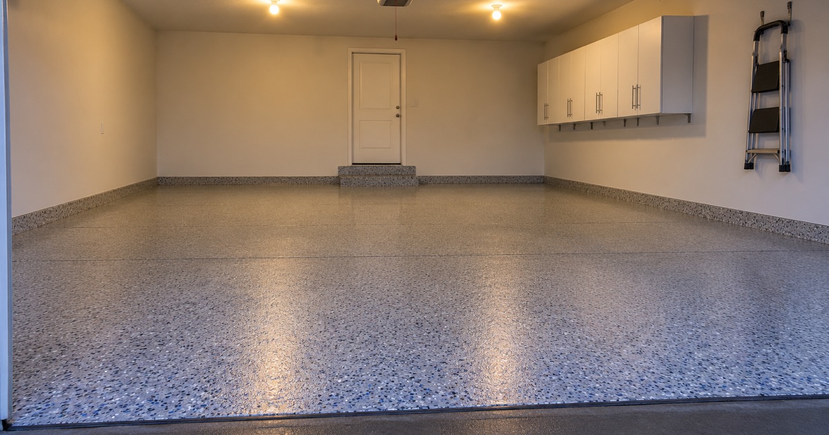An empty, finished gray and blue flake garage floor in a newer Ankeny home at dusk, the overhead door raised and the house door lit.